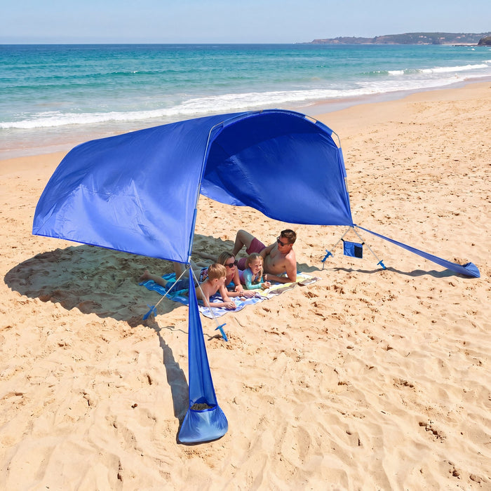 A large blue beach canopy tent is set up on a sandy beach with ocean waves in the background. A family is sitting underneath the canopy on towels, while other beachgoers are visible nearby.